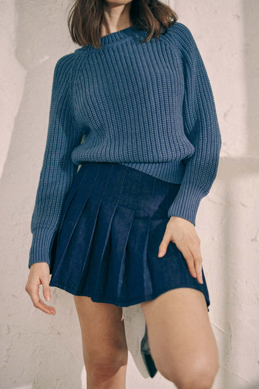 Woman wearing navy denim pleated skirt made from 100% organic cotton, styled with a blue knit sweater.