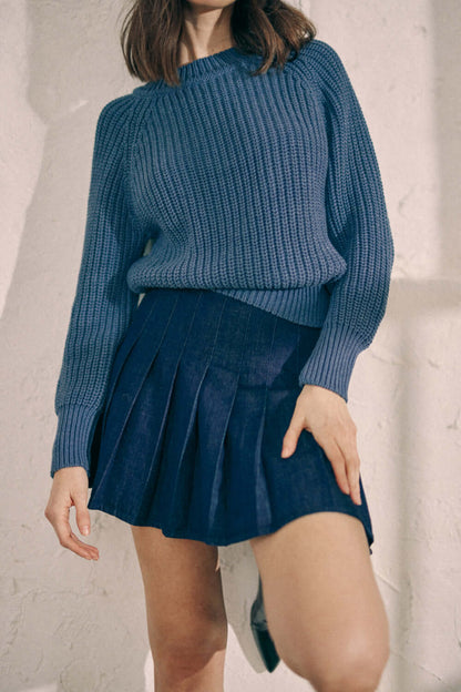 Woman wearing a navy organic cotton sweater and denim pleated skirt, showcasing stylish and ethical fashion.