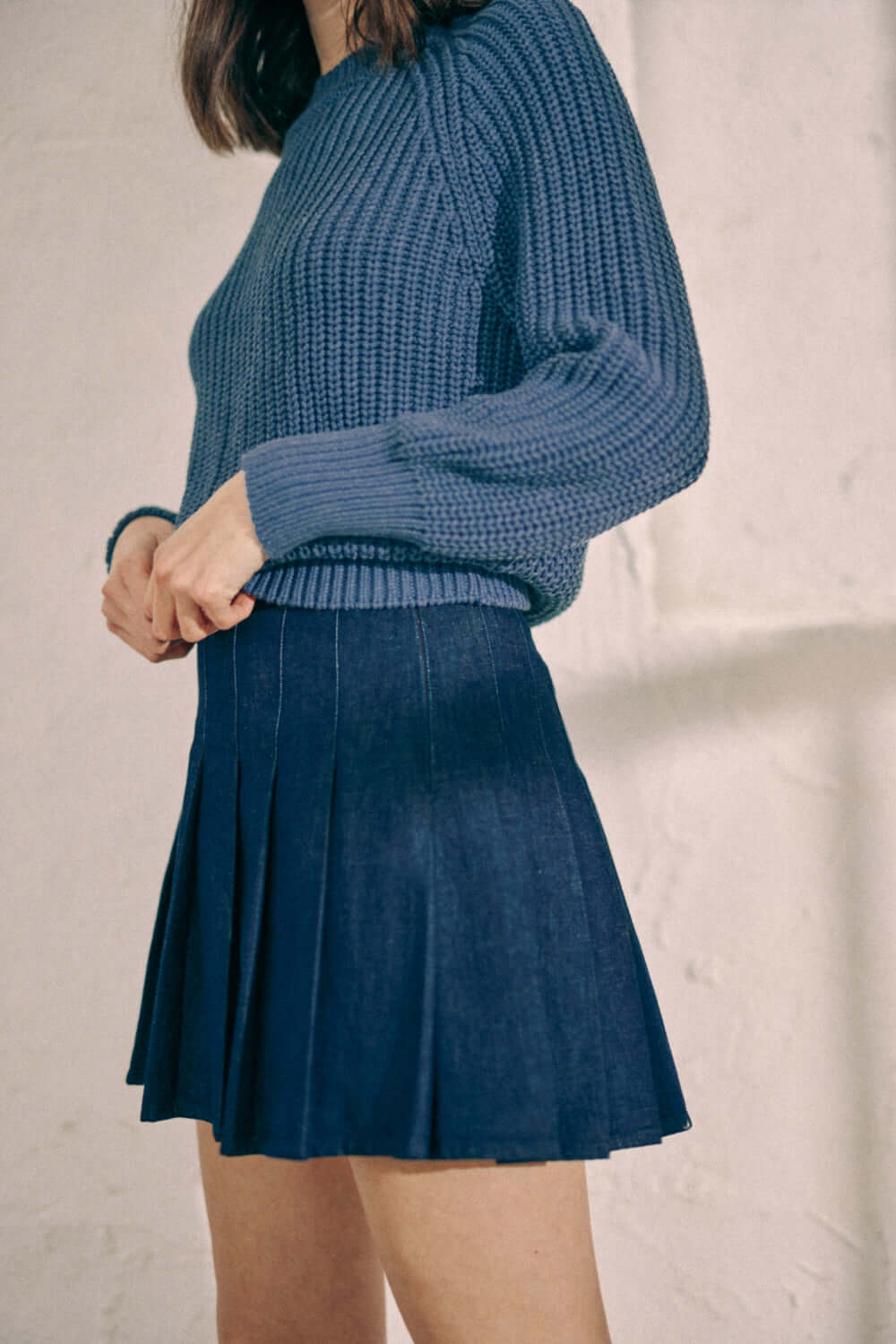 Woman wearing navy denim pleated skirt made from 100% organic cotton, featuring back metal zip, styled with a blue knitted sweater