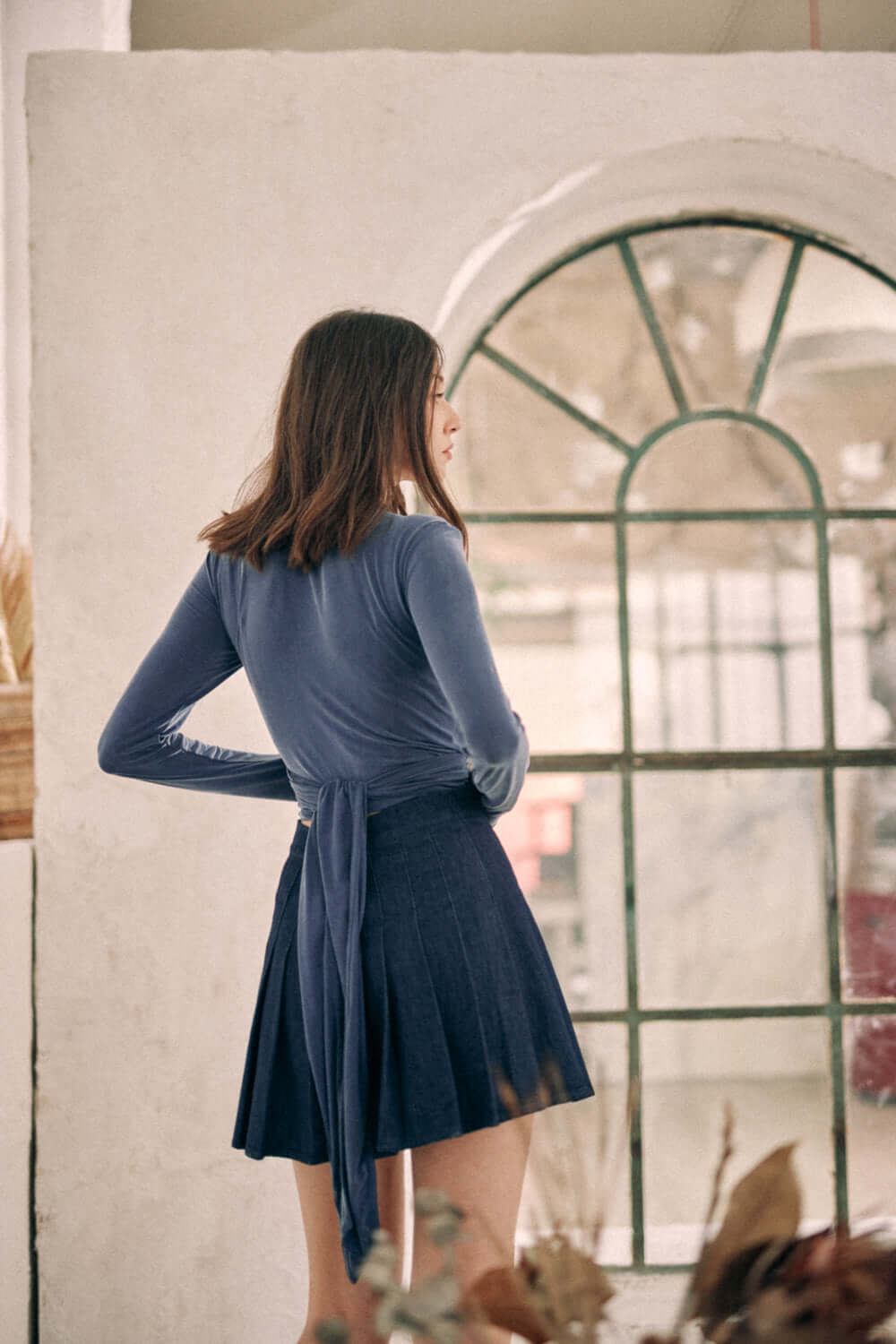 Model wearing navy denim pleated mini skirt made from 100% organic cotton woven denim from Porto, standing near a window.