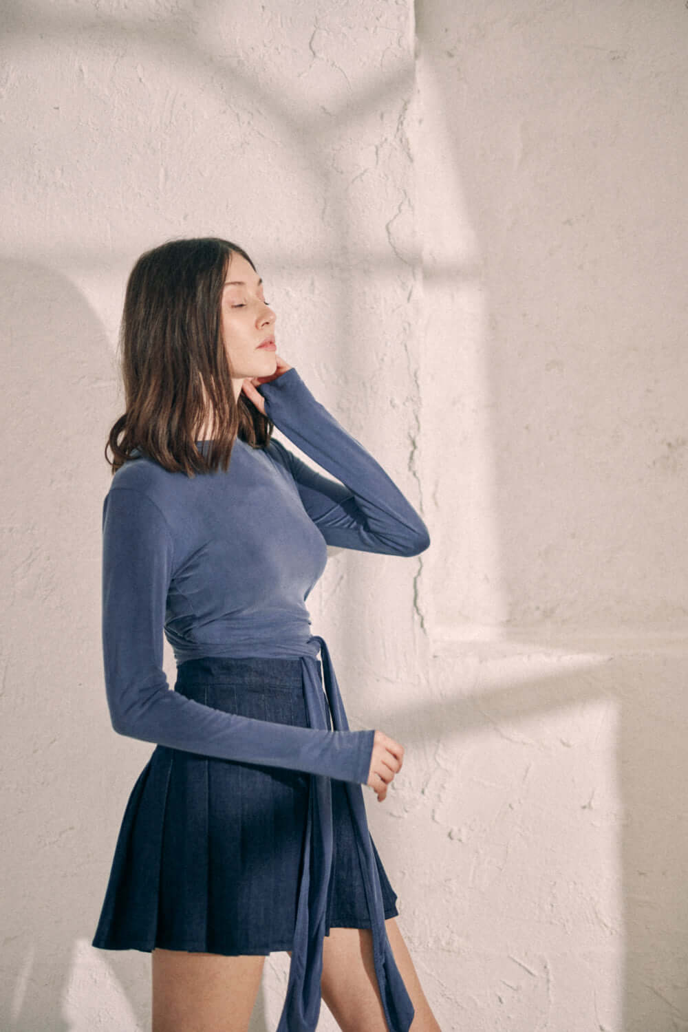 Model wearing NAVY DENIM PLEATED SKIRT made of 100% organic cotton from Porto, styled with a matching long-sleeve top in a bright room.