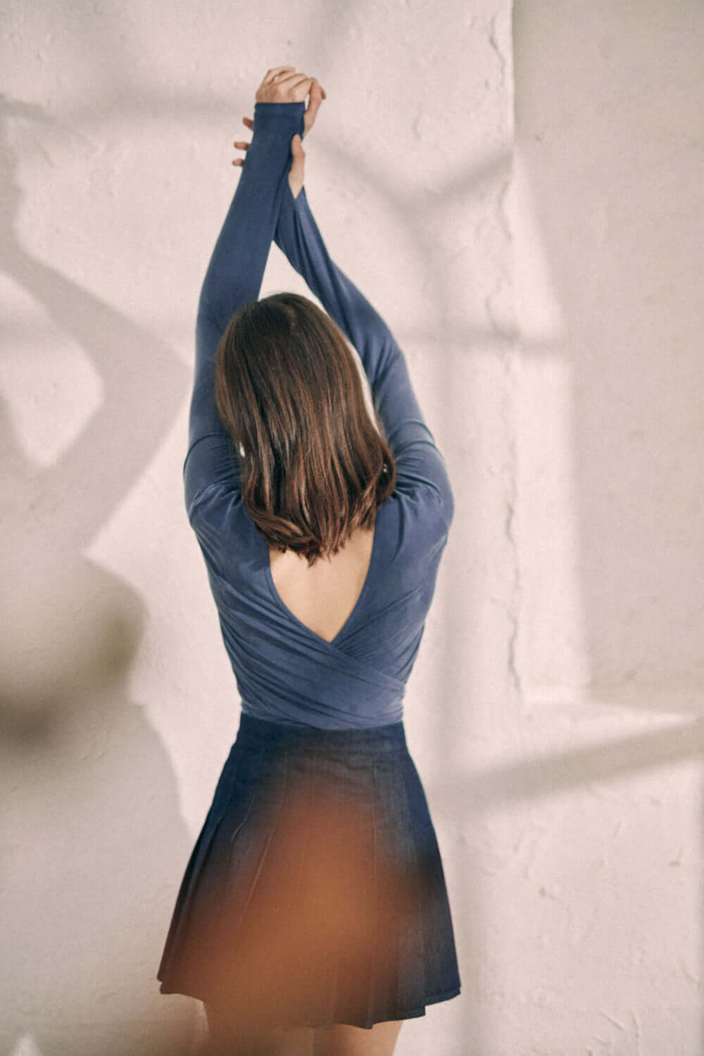 Woman in navy cupro wrap top with thumb holes, seen from the back with arms raised against a light background, wearing a dark skirt