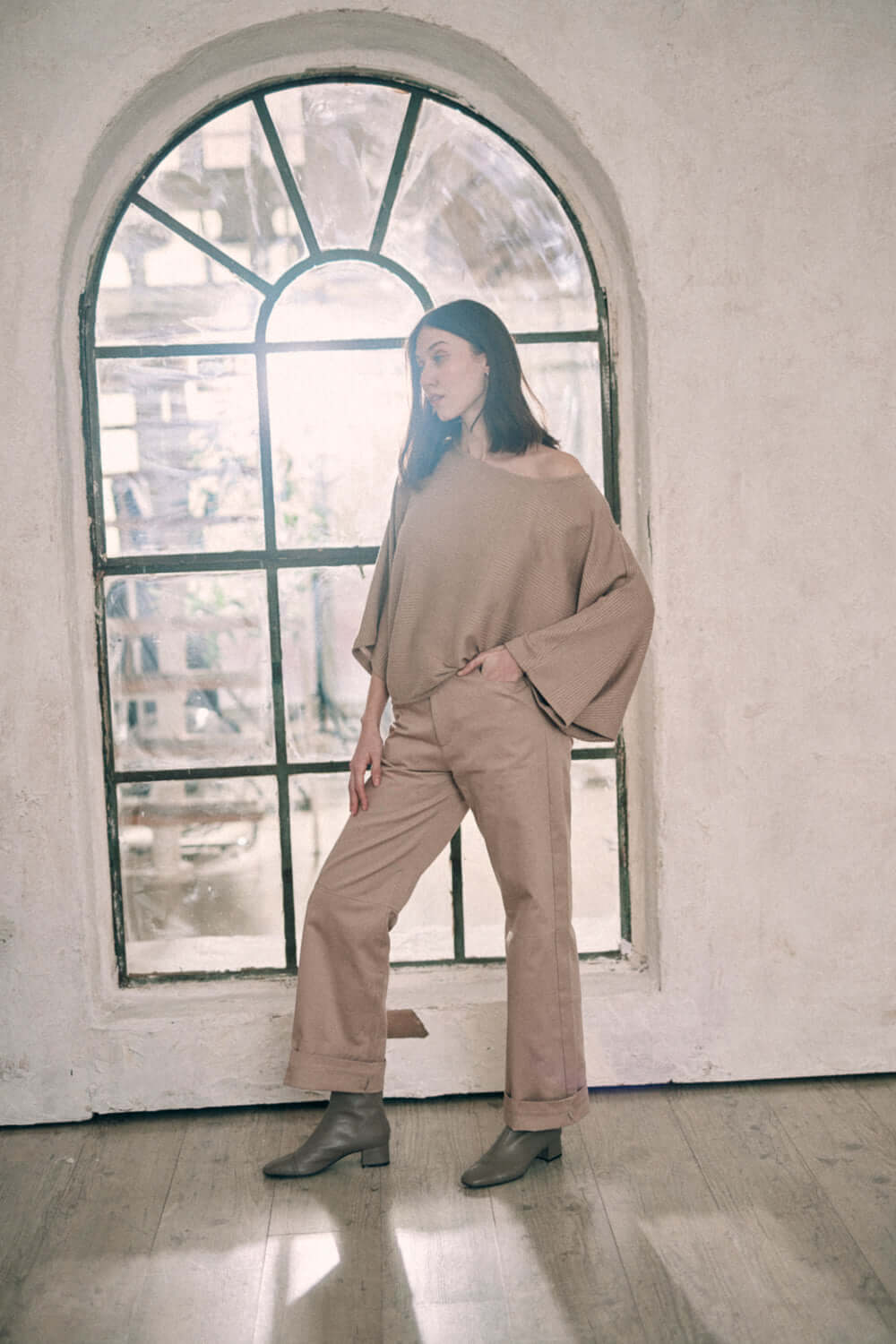 Model wearing taupe batwing top and trousers by window, crafted from recycled polyester and organic cotton jersey, sustainable fashion