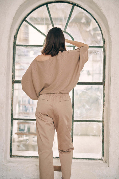 Woman wearing taupe denim trousers with turned ankles and an off-shoulder sweater standing by a large arched window.