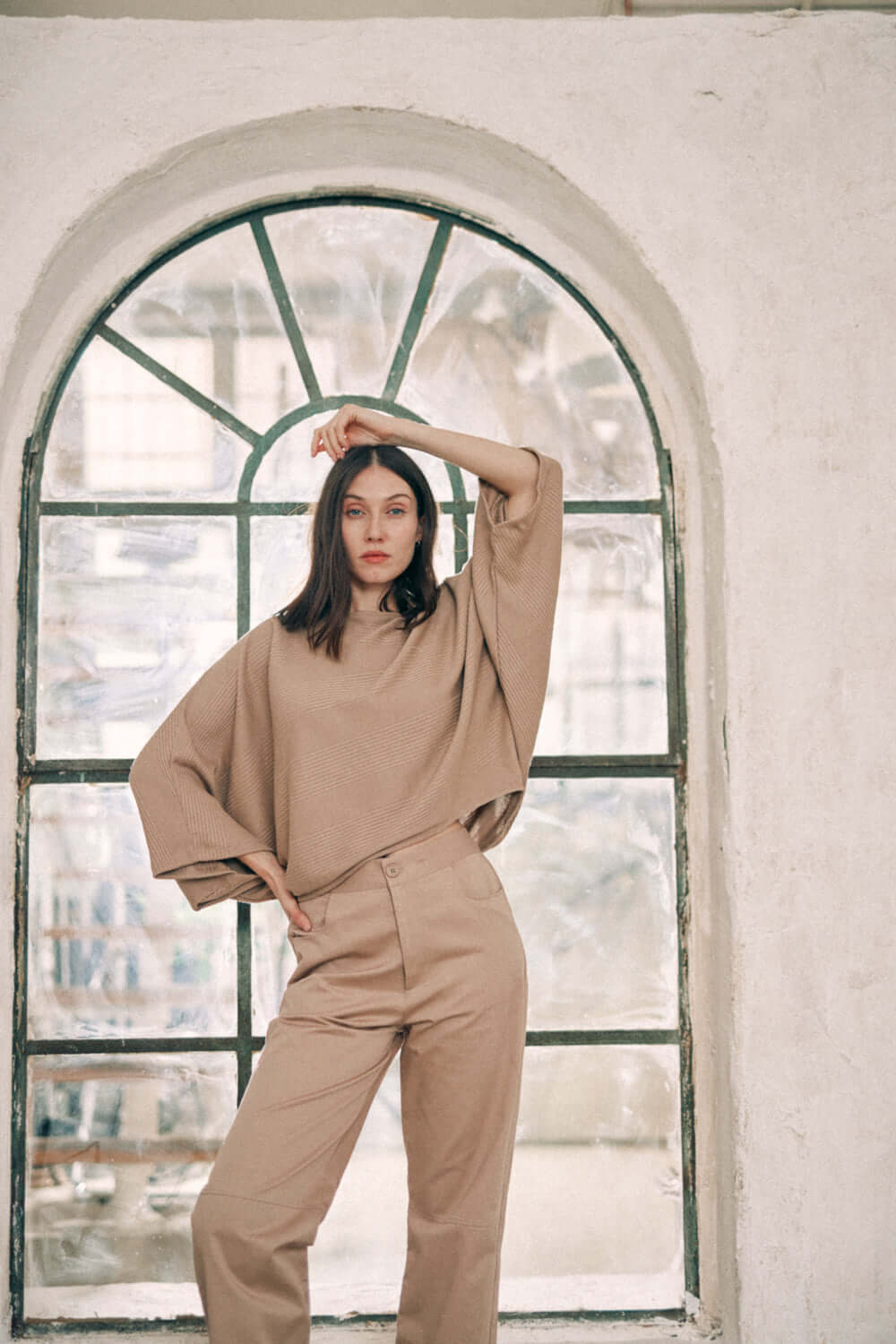 Woman wearing taupe batwing top and trousers in front of an arch window, showcasing a fashion-forward outfit made with recycled and organic materials.