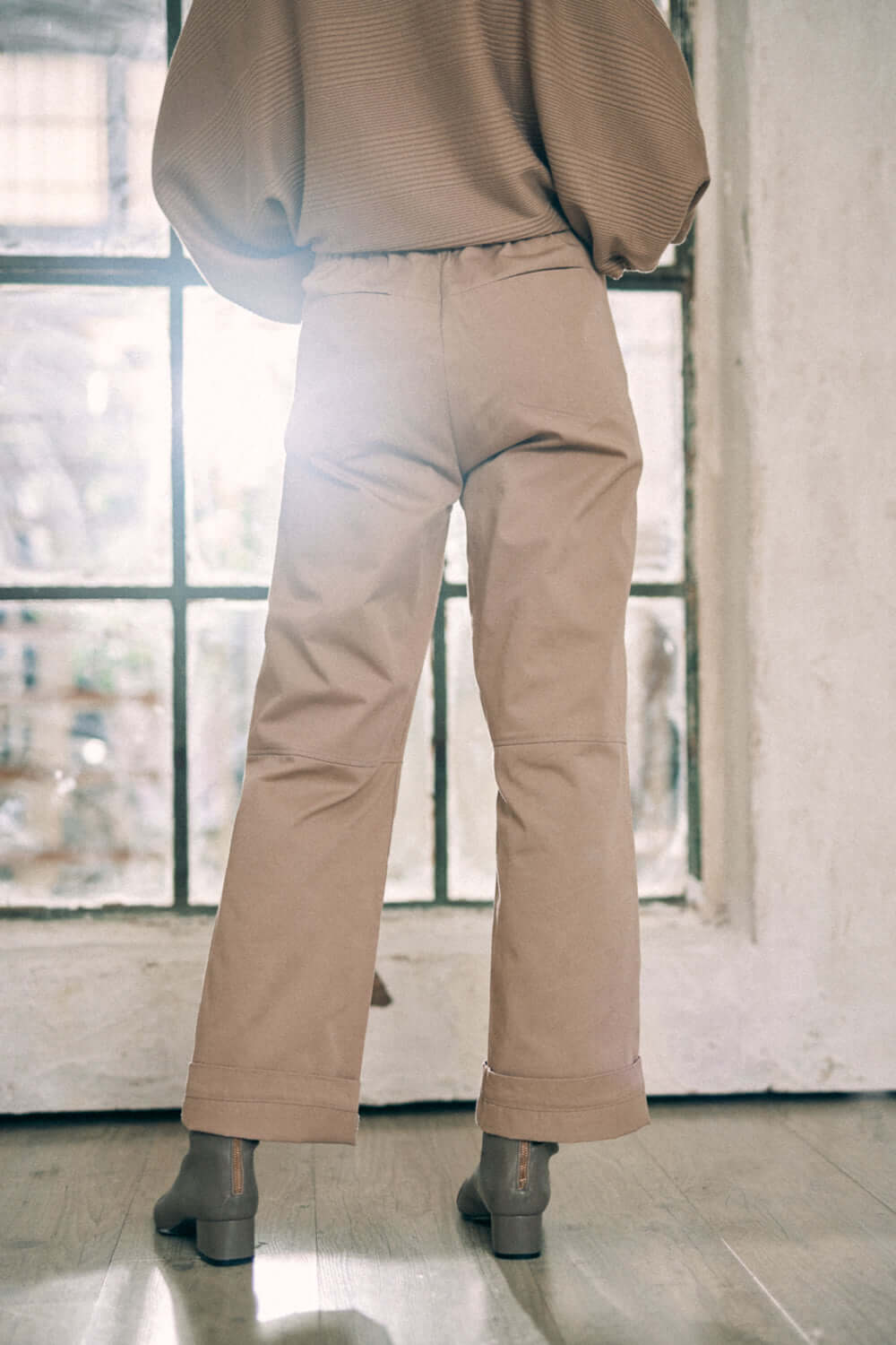 Back view of model wearing taupe denim trousers made from 100% organic cotton, showcasing turned ankles and branded COSSAC buttons.