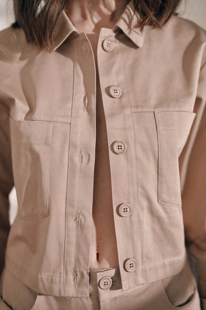 Close-up of taupe denim jacket featuring customised buttons, made from 100% organic cotton, worn by model with turned sleeves.