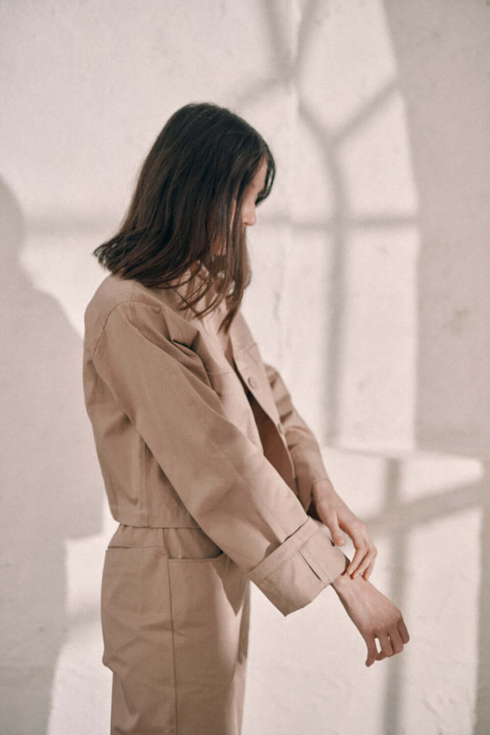 Woman wearing taupe denim jacket made of 100% organic cotton, adjusting sleeves in a bright room.