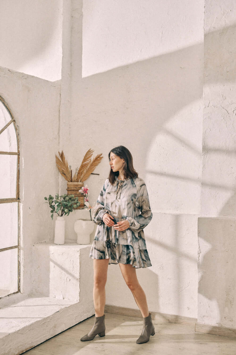 Woman wearing printed mini dress with flowy fit and frills, made from 100% Ecovero viscose in autumn print, standing in bright room.