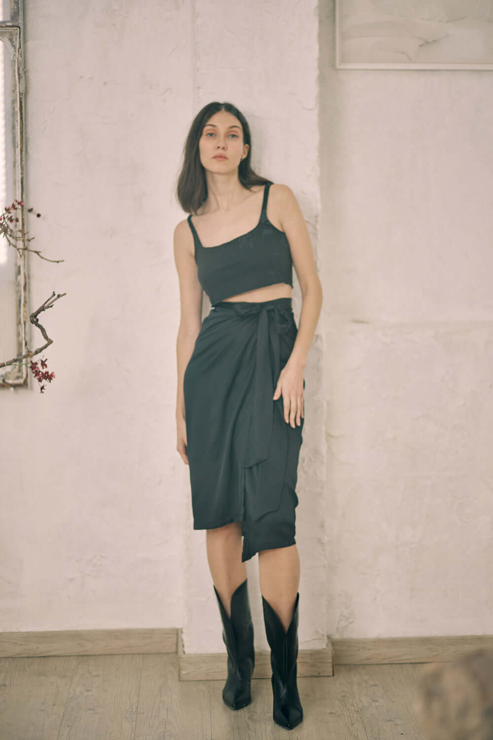 Model wearing a black wrap midi skirt styled with a crop top and knee-high boots in a minimalist setting.
