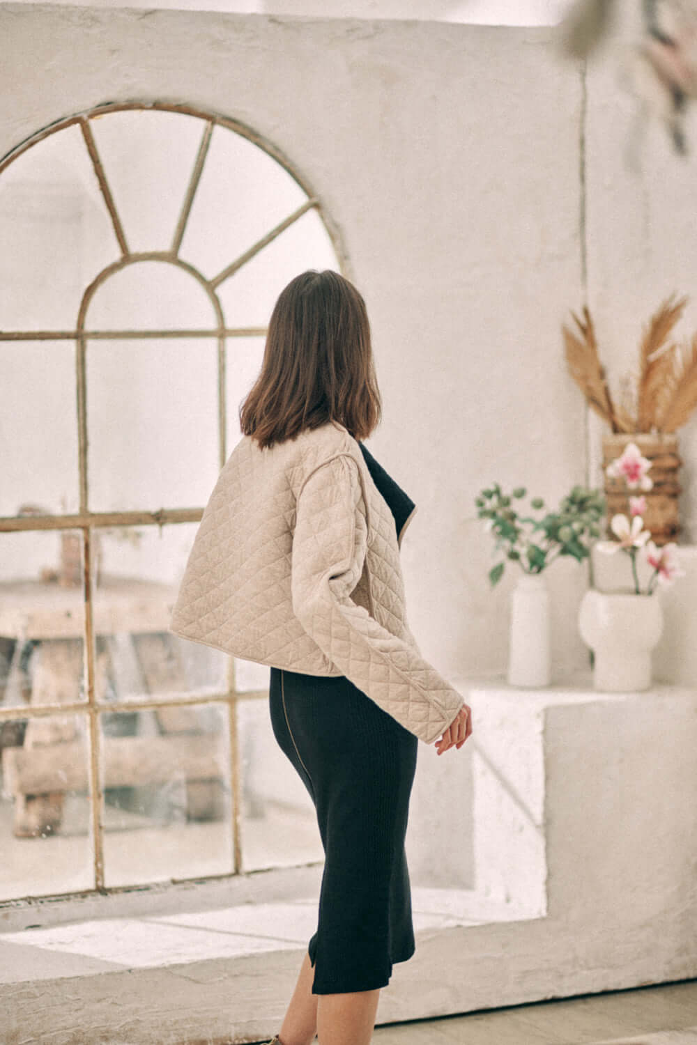 Woman wearing a reversible padded jacket in beige, made from organic cotton and recycled polyester, standing in front of a rustic window.