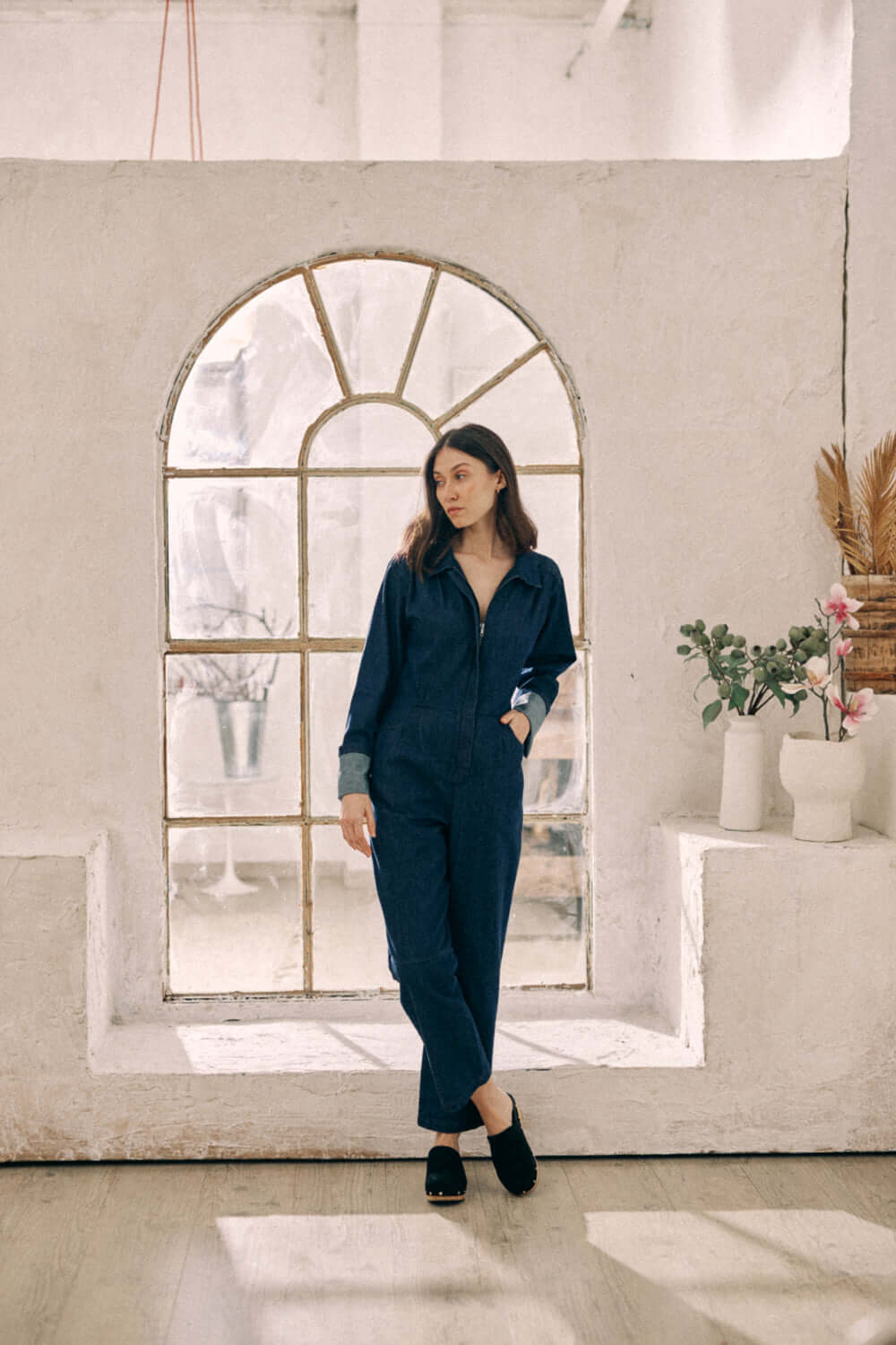 Woman wearing navy denim boilersuit made from organic cotton, standing by a window with turned cuffs, showcasing timeless style.