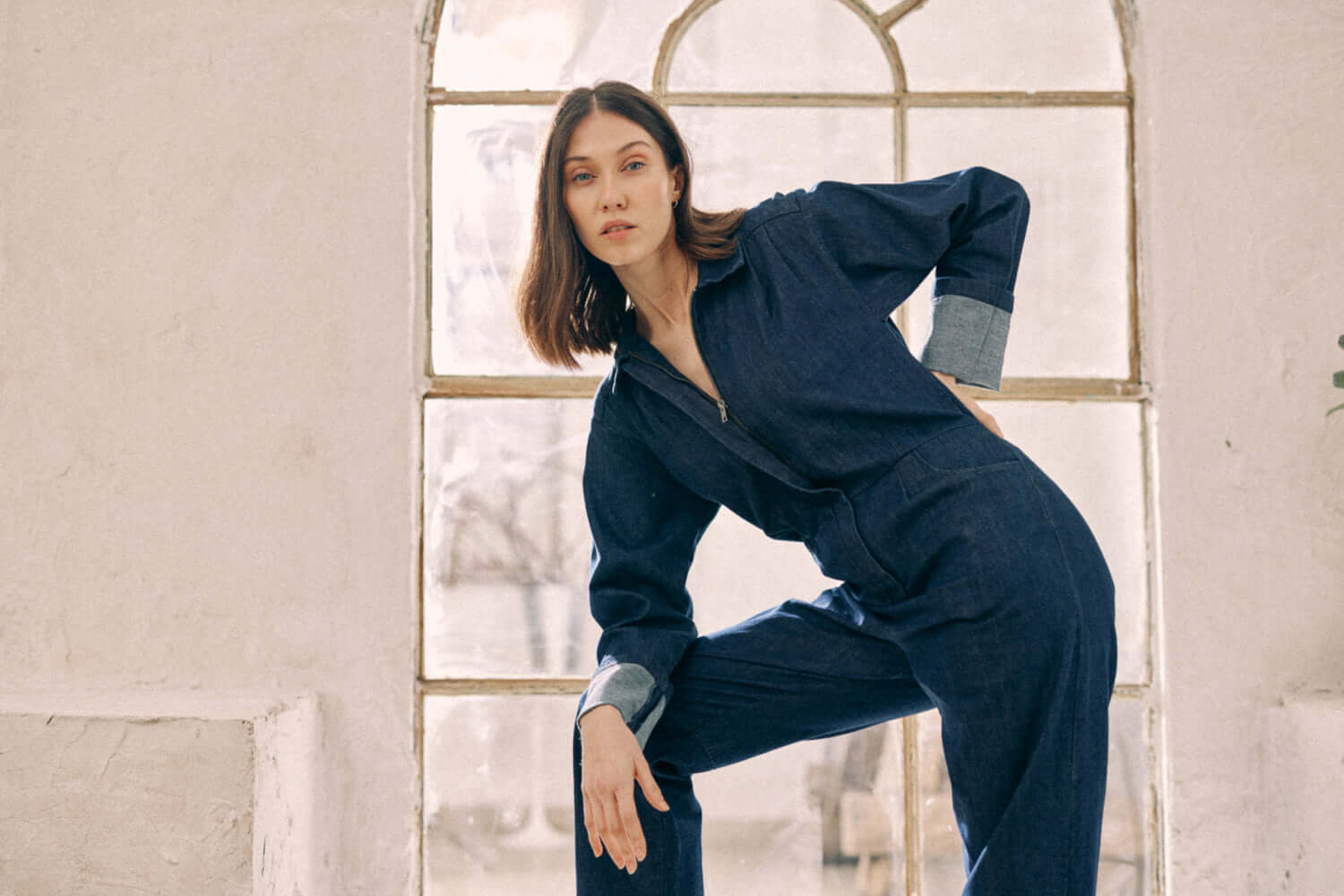 Woman wearing navy denim boilersuit with rolled cuffs, posing elegantly in front of arched window, made from organic cotton.