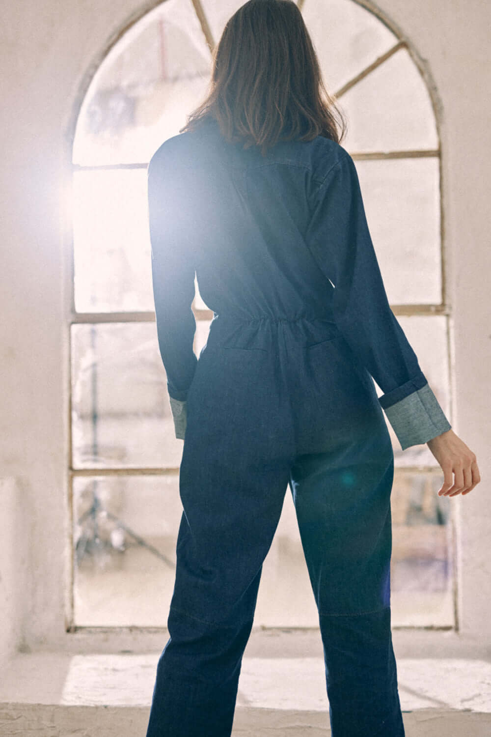 Woman wearing a navy denim boilersuit with cuffs turned up, standing in front of a window.