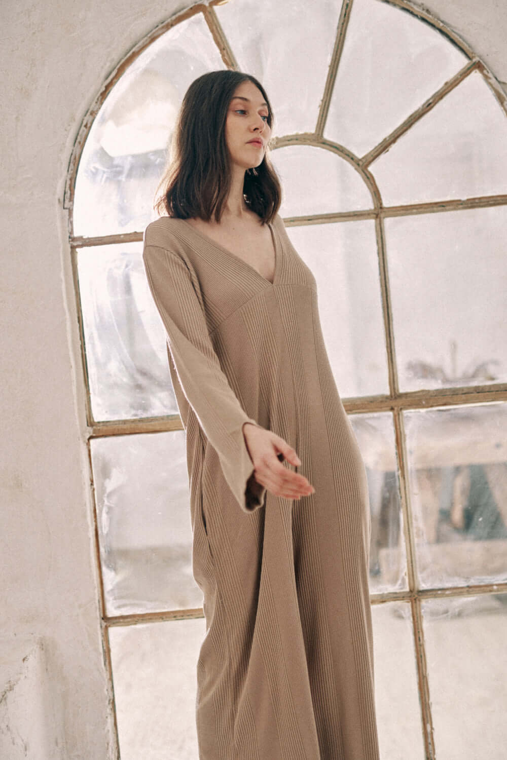 Model wearing a taupe wide jumpsuit made from 40% recycled polyester and 60% organic cotton, standing by a window.