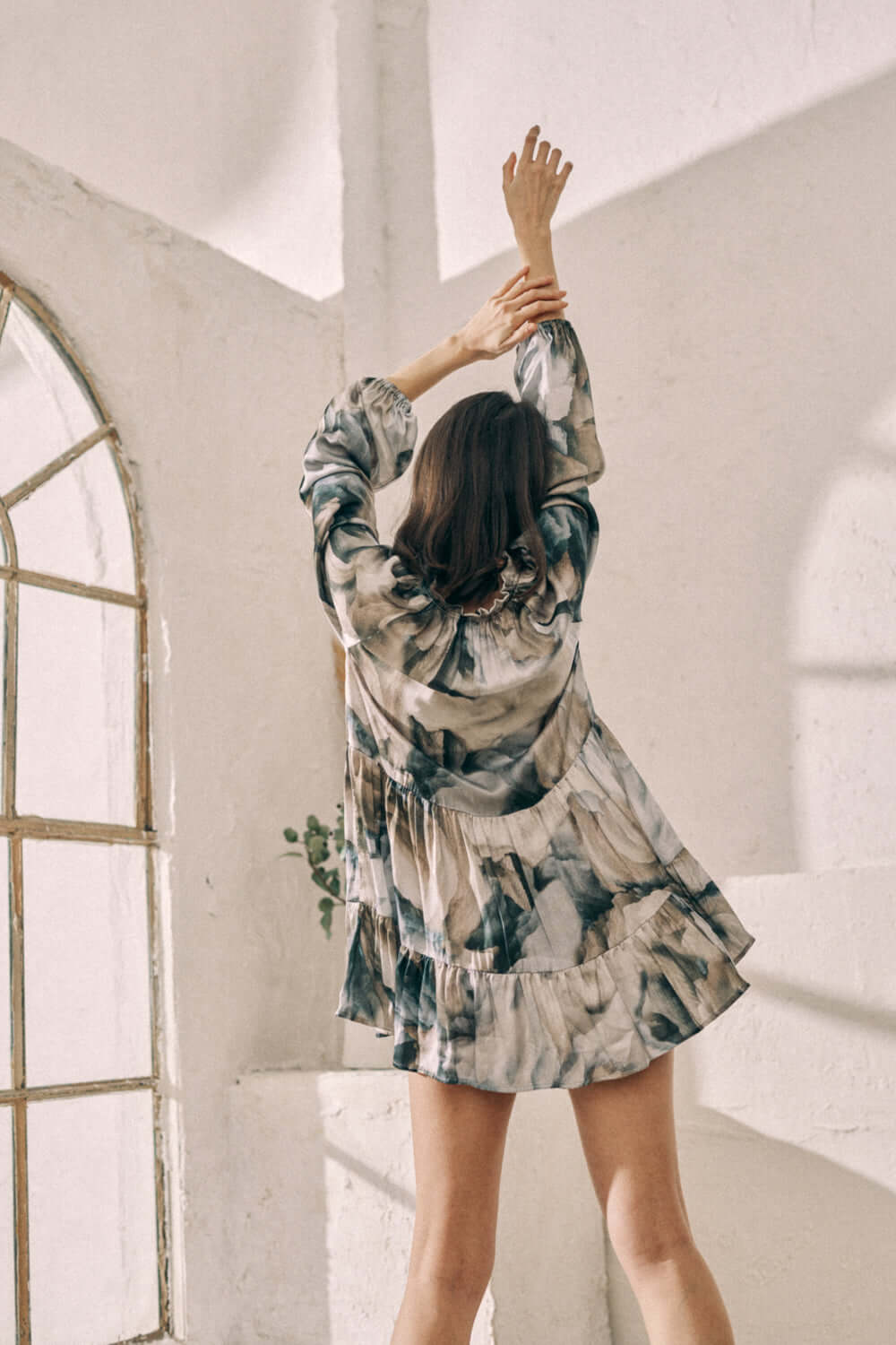 Woman wearing printed mini dress with Ecovero viscose, showcasing flowy fit and frills, posing in bright room with vintage window.