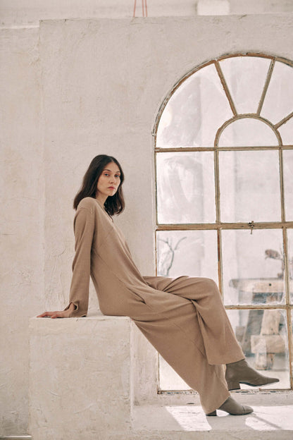 Woman wearing taupe wide jumpsuit made of thick jersey fabric, sitting indoors beside an arched window.