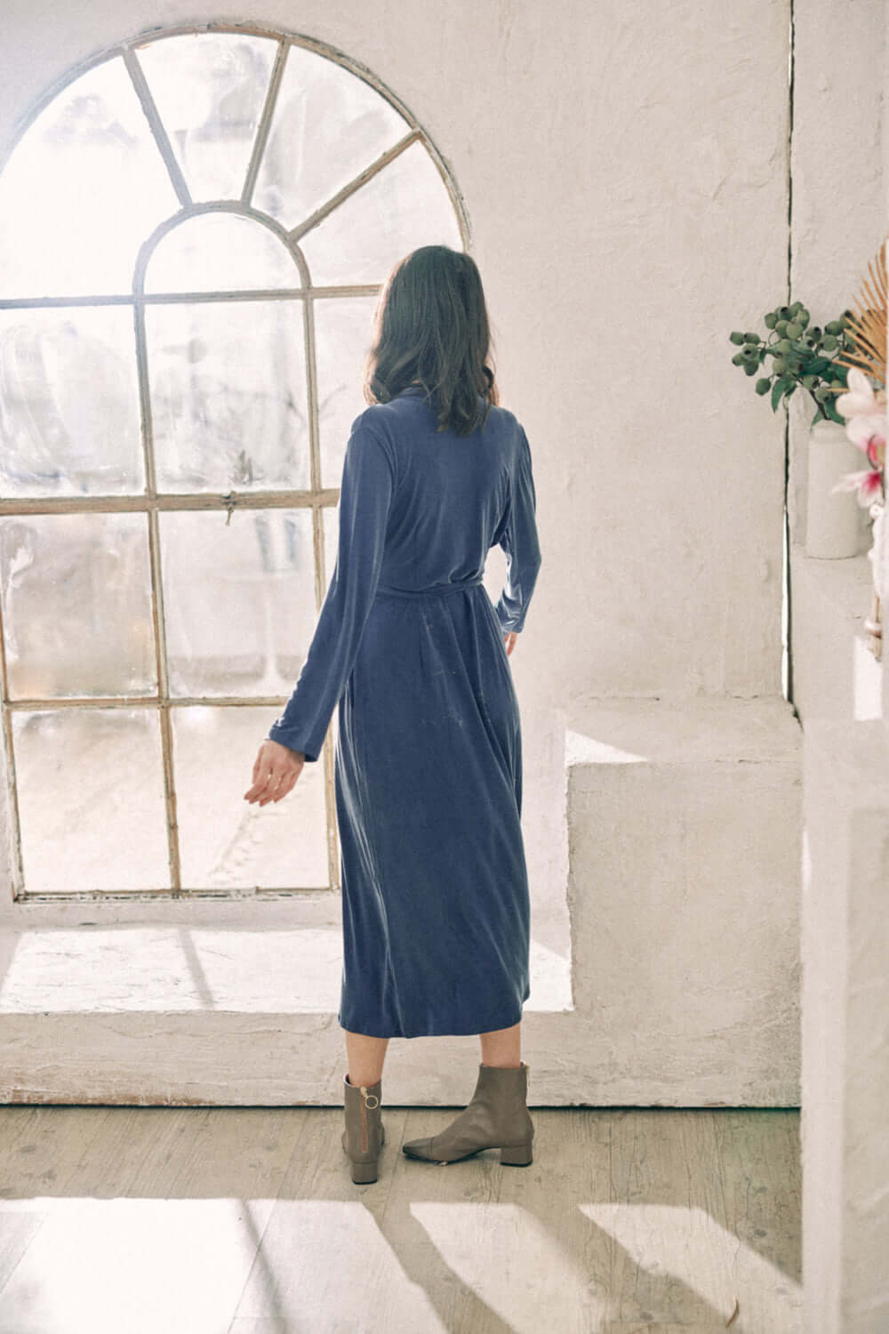 Woman wearing navy cupro wrap dress with velvety finish in front of vintage window in bright room.