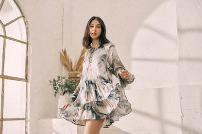 Woman wearing Ecovero printed mini dress with flowy fit and frills, standing in a bright room next to a window and plants.