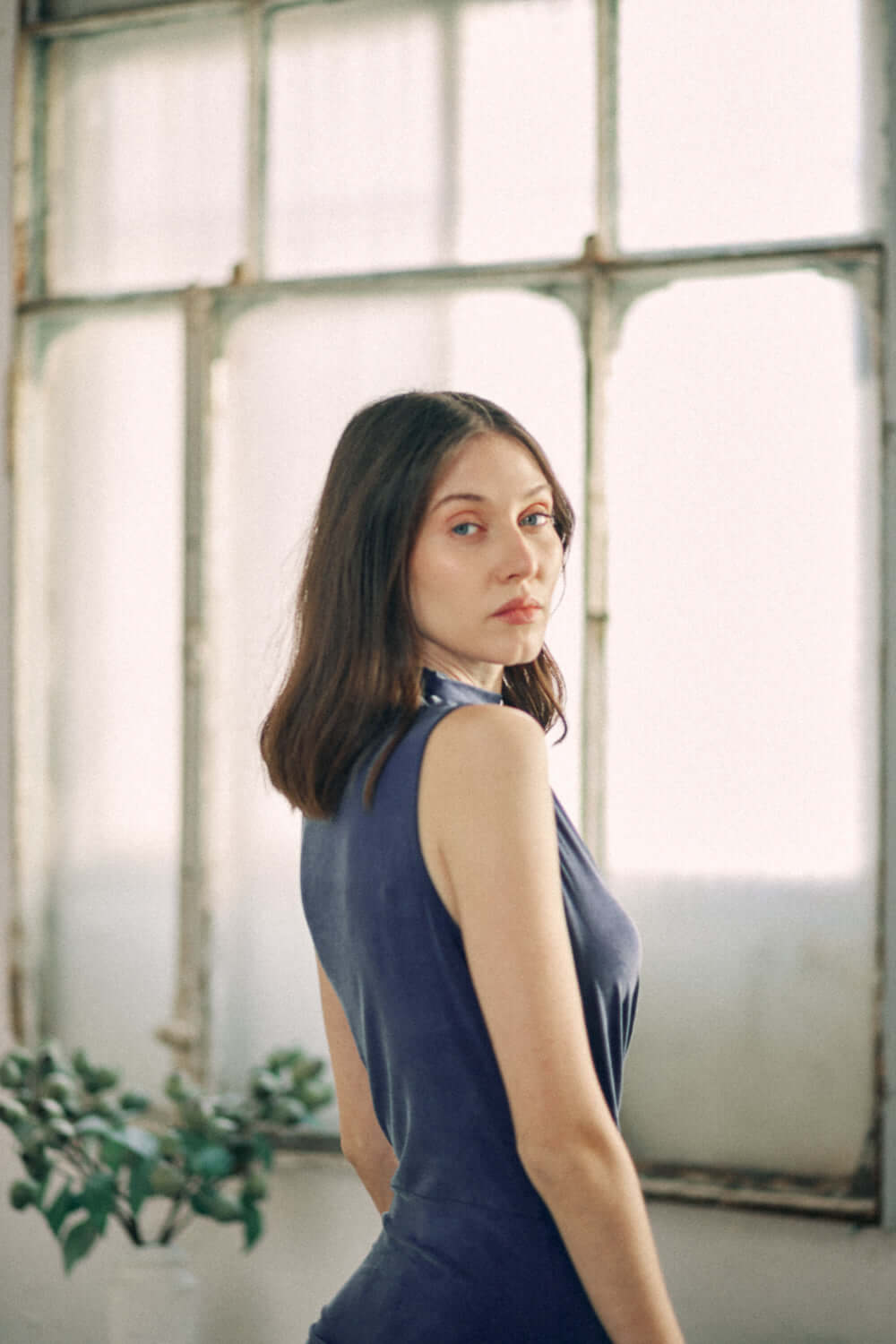 Woman wearing navy cupro bodysuit with drape detail, standing by a window.