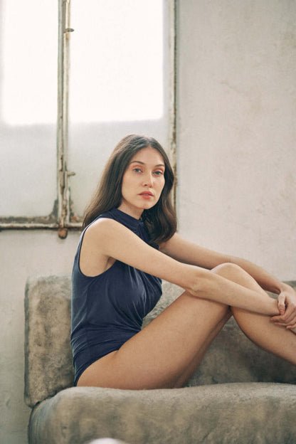 Woman wearing navy cupro bodysuit with velvety finish and drape detail bust, seated on a cushioned surface. Model in stylish bodysuit.