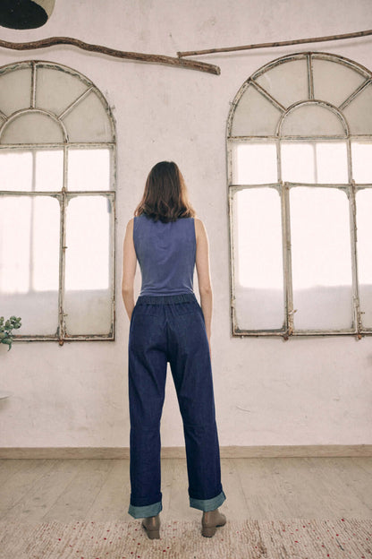 Woman wearing navy denim trousers made from 100% organic cotton, shown from the back with ankles turned up, stands in front of vintage windows.