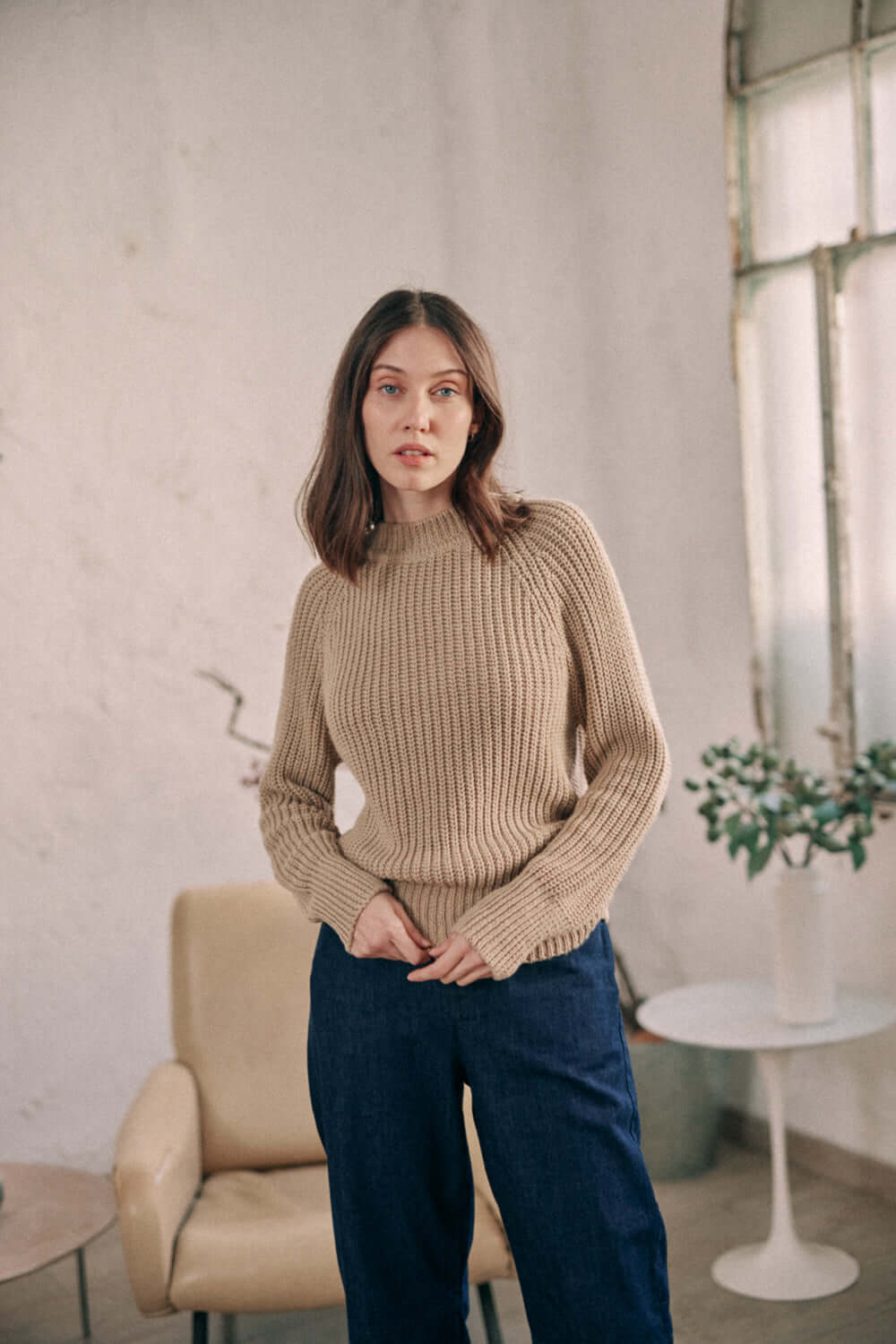 Woman wearing taupe sweater made of vegan organic cotton, standing in a cozy indoor setting. Exclusive online collection.