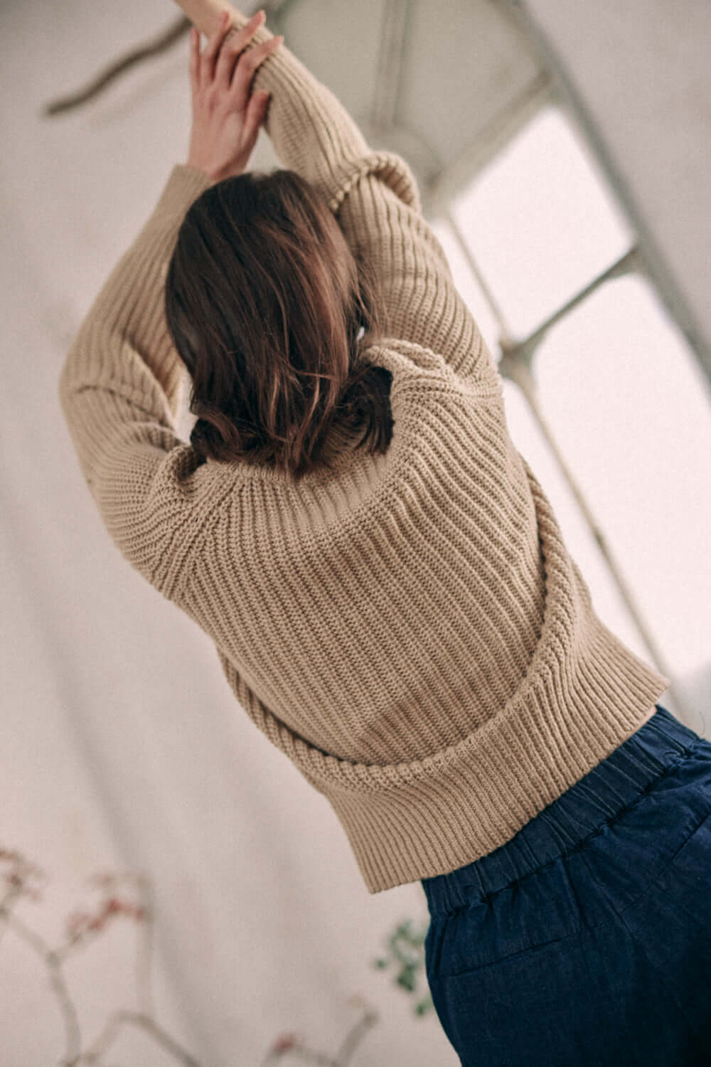 Woman wearing taupe sweater made of organic cotton, showcasing ribbed texture and crew neck, posed in a bright room.