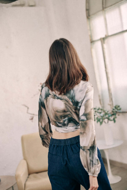 Woman wearing autumn print wrap top in Ecovero viscose, shown from the back, paired with blue pants, standing in a cozy room.