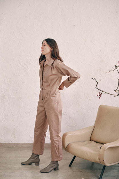 Woman wearing taupe denim boilersuit made from organic cotton, modeling next to a beige chair with ankles turned up.