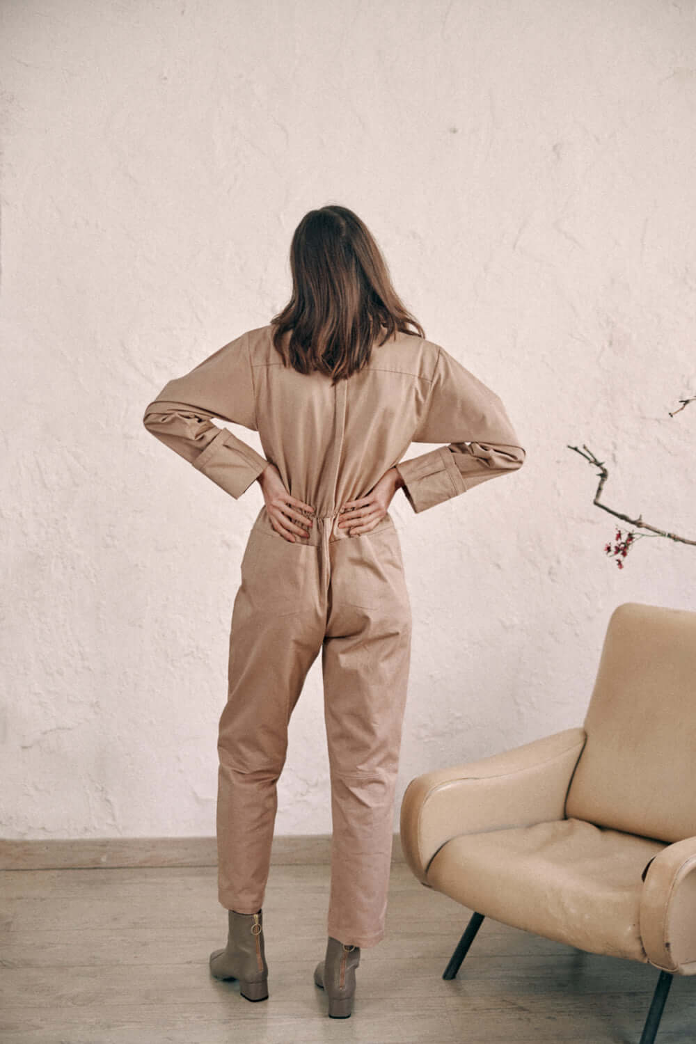 Woman wearing taupe denim boilersuit made of 100% organic cotton, standing back-to-camera next to beige chair.