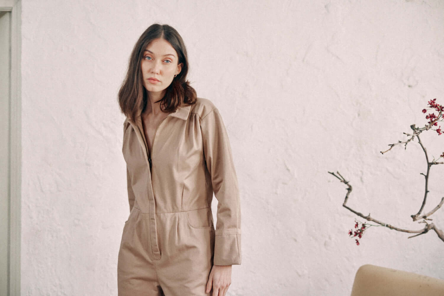 Woman wearing taupe denim boilersuit made from organic cotton, standing against a white wall with a minimalist decor.