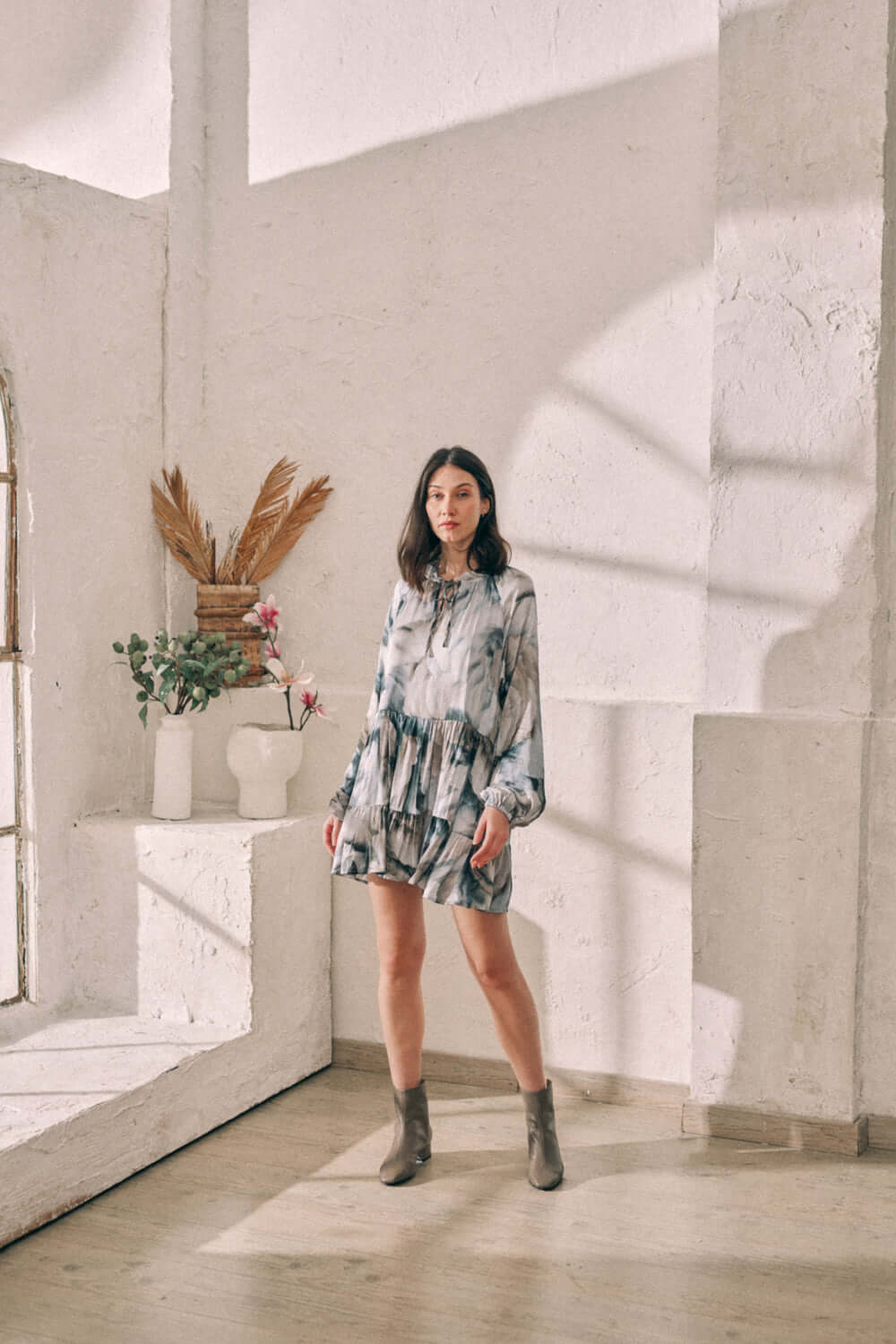 Woman wearing flowy mini dress with Ecovero autumn print, standing in bright room with natural light and decorative plants.