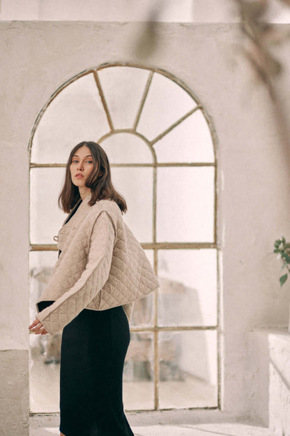 Woman wearing a beige reversible padded jacket made from organic cotton and recycled polyester, in a minimalist setting.