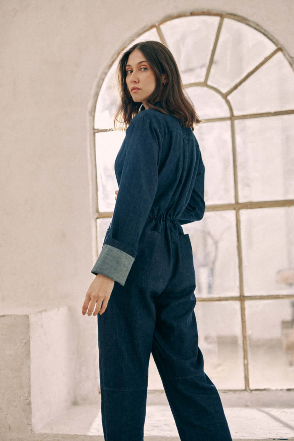 Woman wearing navy denim boilersuit in front of arched window, showcasing cuffed sleeves and relaxed fit. Made from organic cotton.