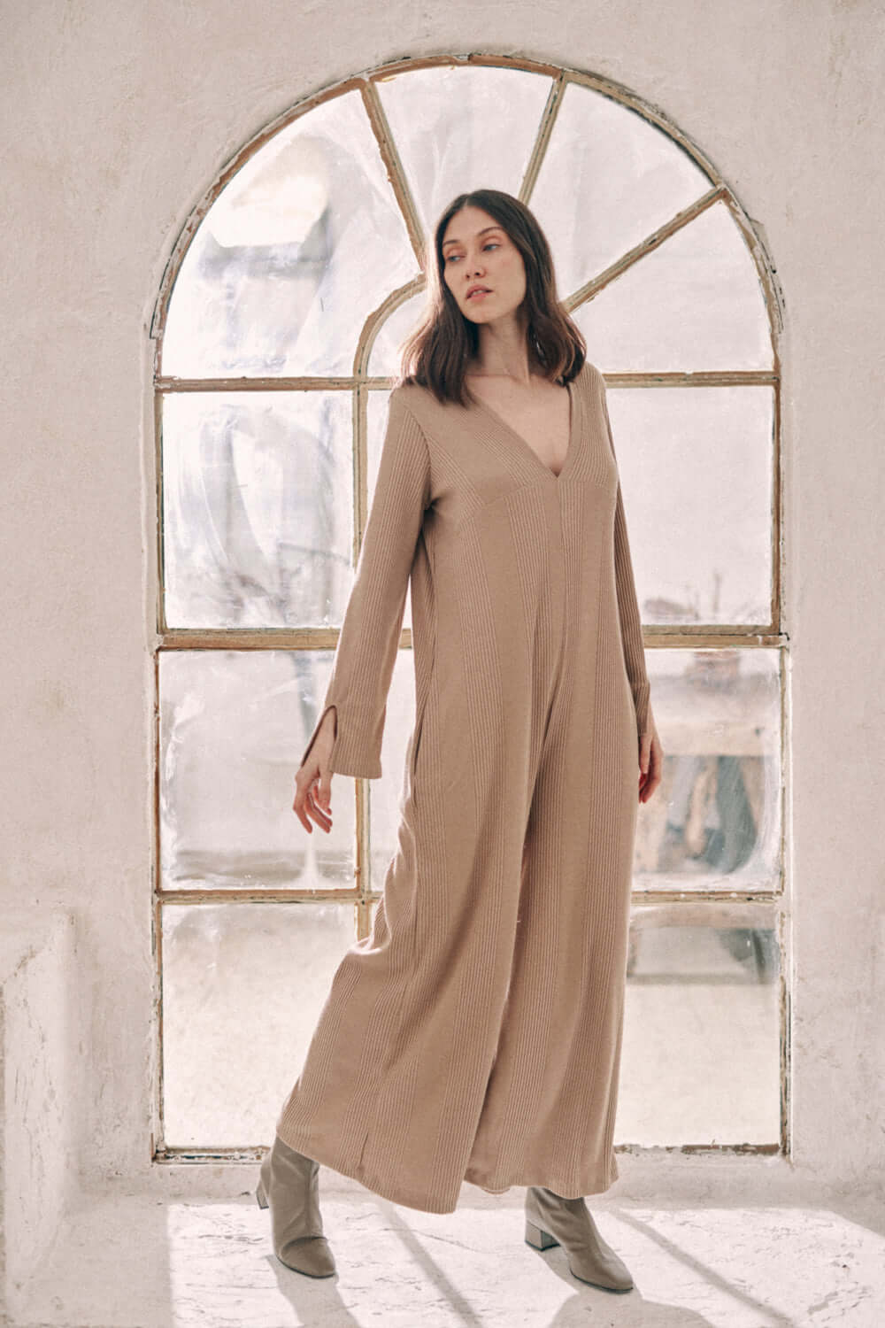 Woman wearing taupe wide jumpsuit crafted from thick jersey fabric, standing in front of vintage window.