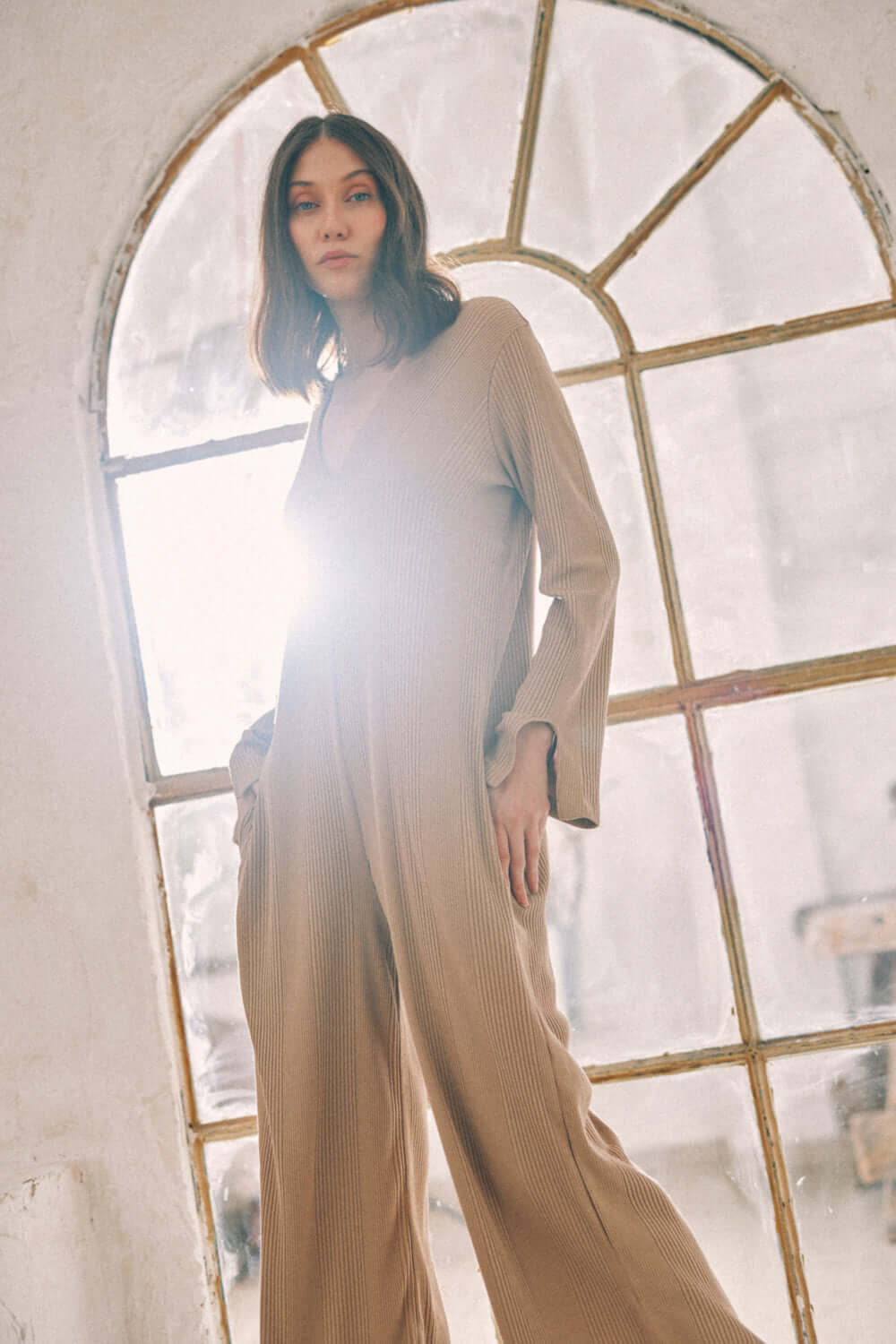 Woman wearing taupe wide jumpsuit crafted in thick jersey fabric standing in front of arched window.