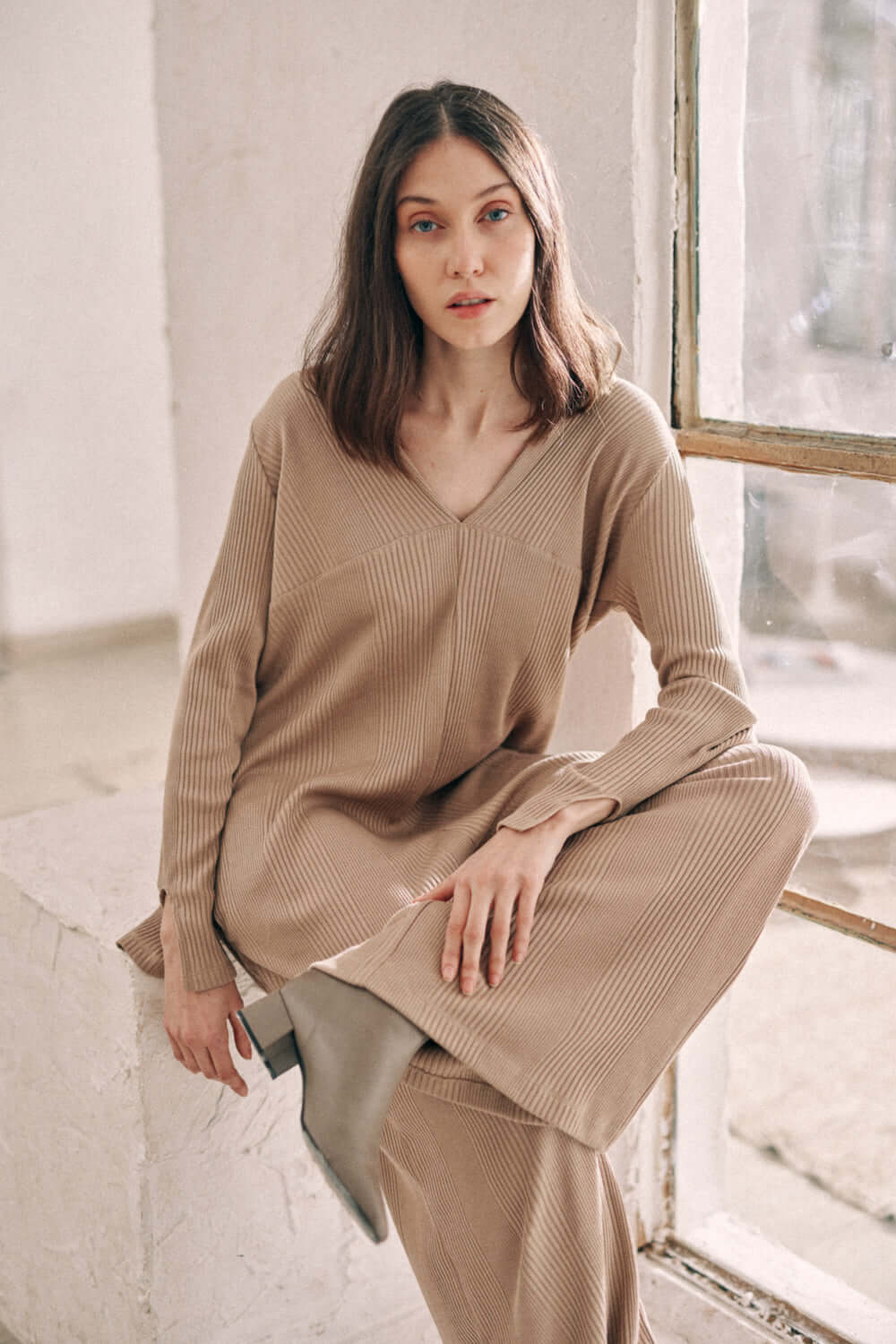 Woman wearing taupe wide jumpsuit made from thick jersey fabric, sitting by a window.