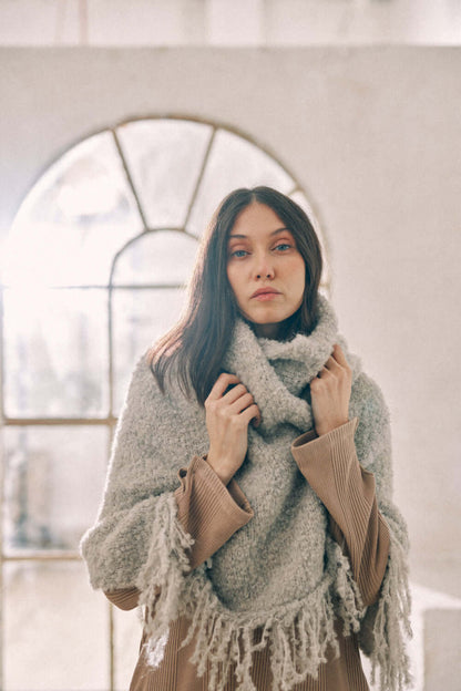 Woman wearing a versatile grey poncho scarf made from sustainable alpaca and recycled polyester, styled in a cozy indoor setting.