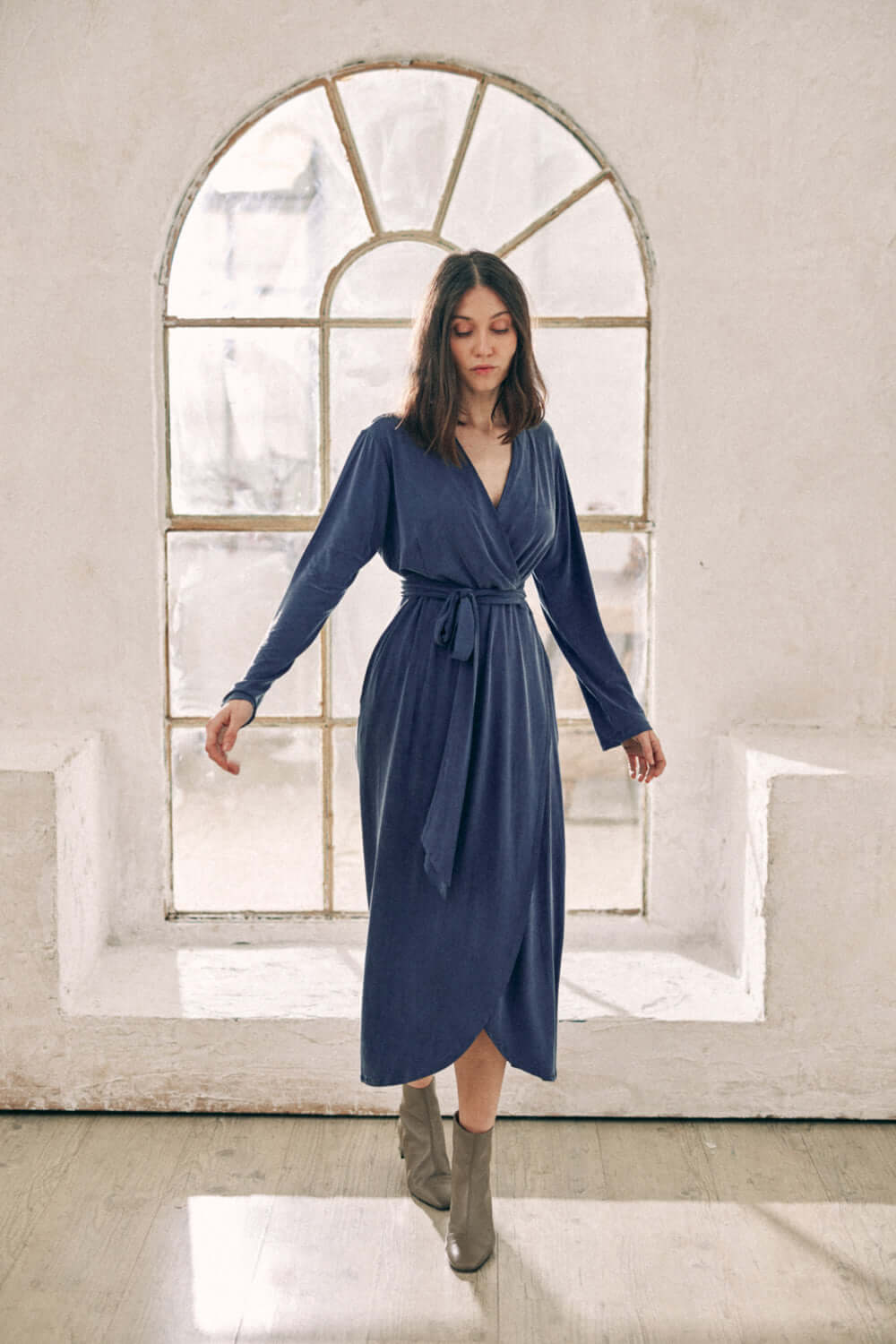 Woman wearing navy cupro wrap dress, midi length with velvety finish, standing in front of an arched window.