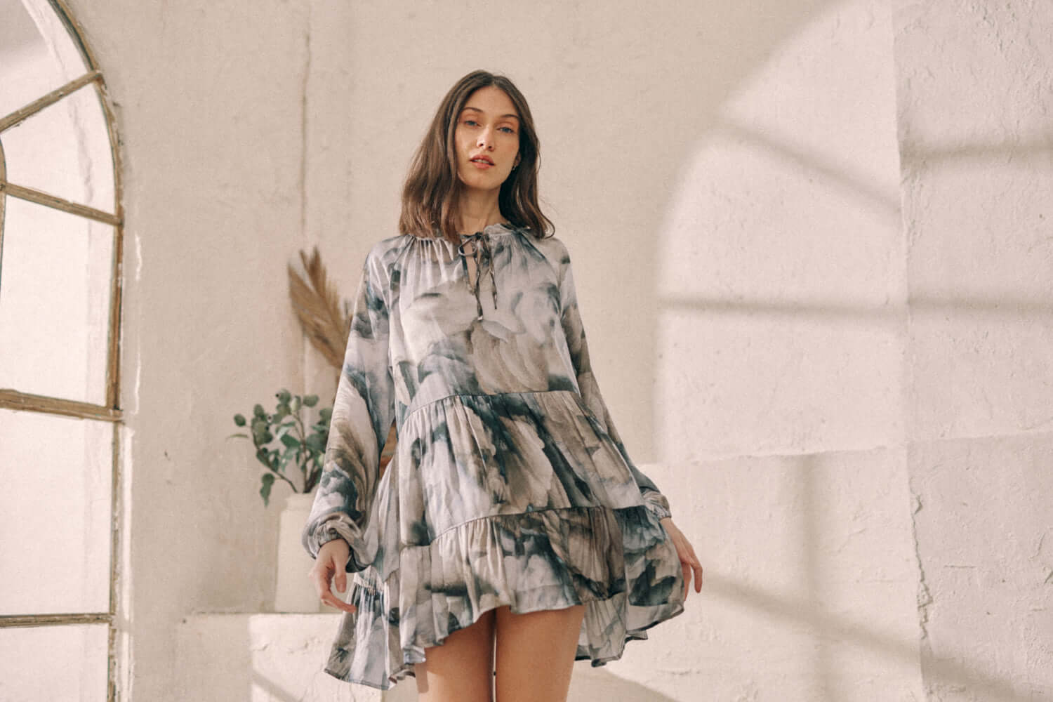 Woman wearing flowy printed mini dress with frills and autumn Ecovero fabric, standing in sunlit room.