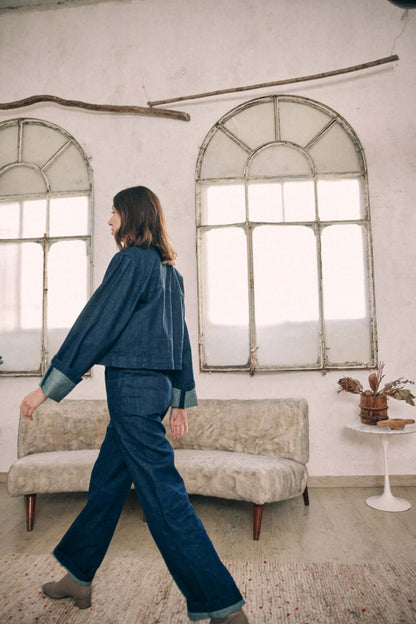 Woman wearing navy denim trousers and jacket, standing indoors near a rustic window.