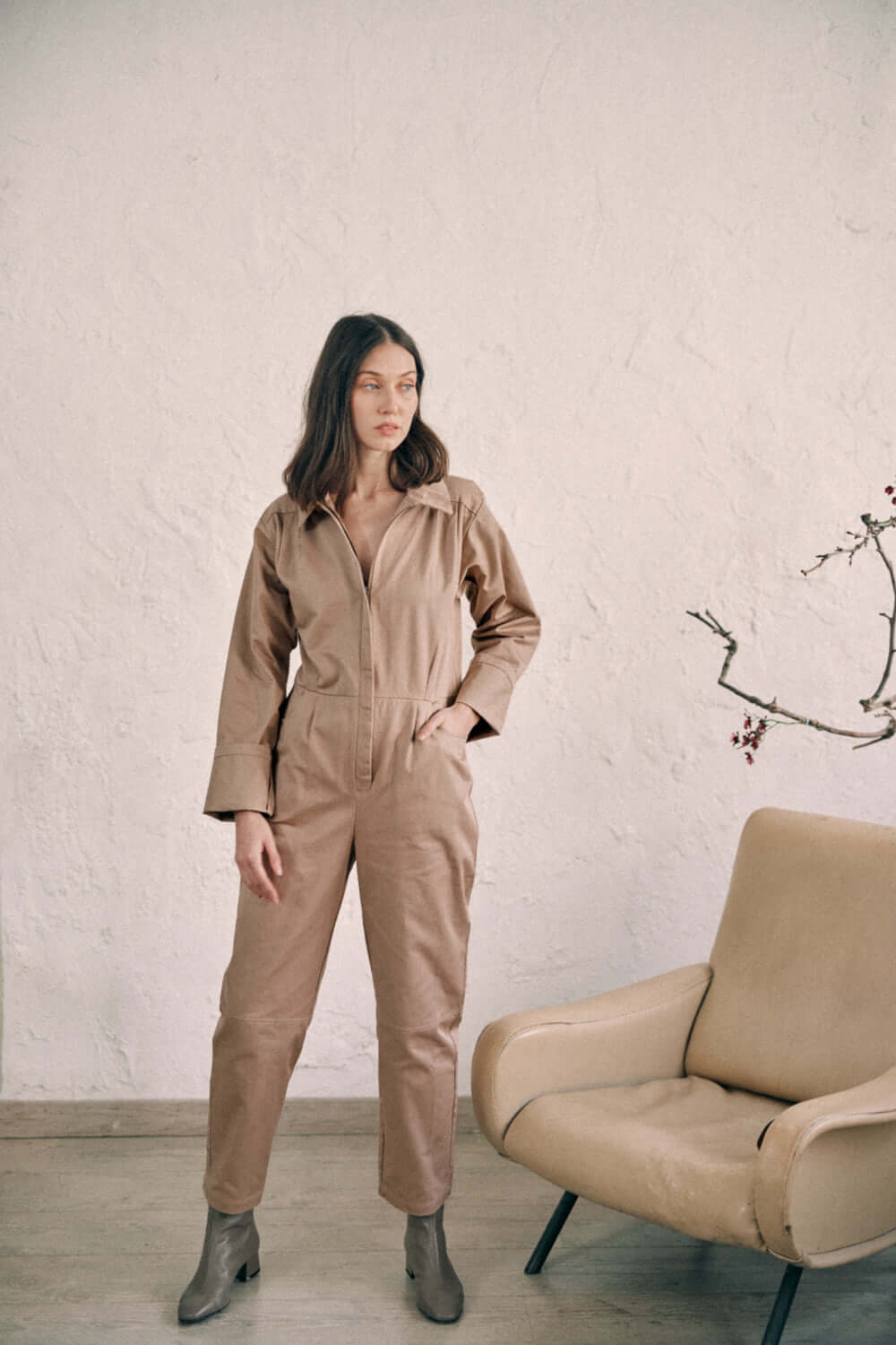 Woman wearing taupe denim boilersuit made from organic cotton, standing beside a beige chair, showcasing timeless fashion style.