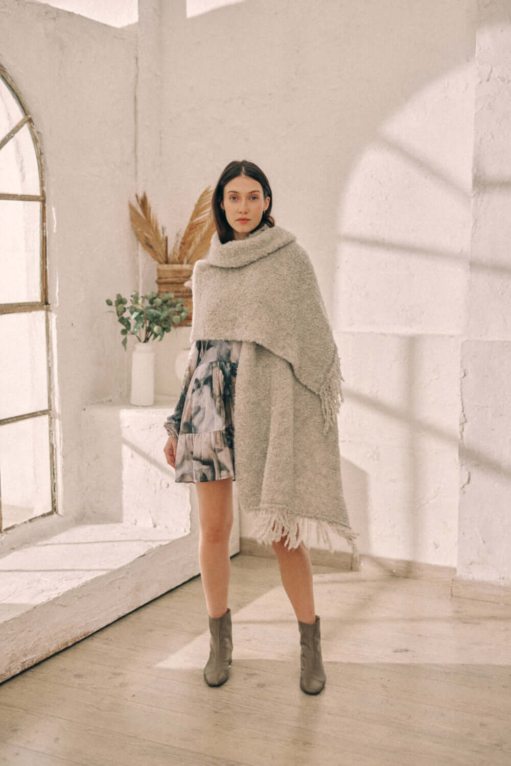 Woman modeling versatile grey poncho scarf, eco-friendly alpaca and recycled polyester, in a stylish indoor setting.