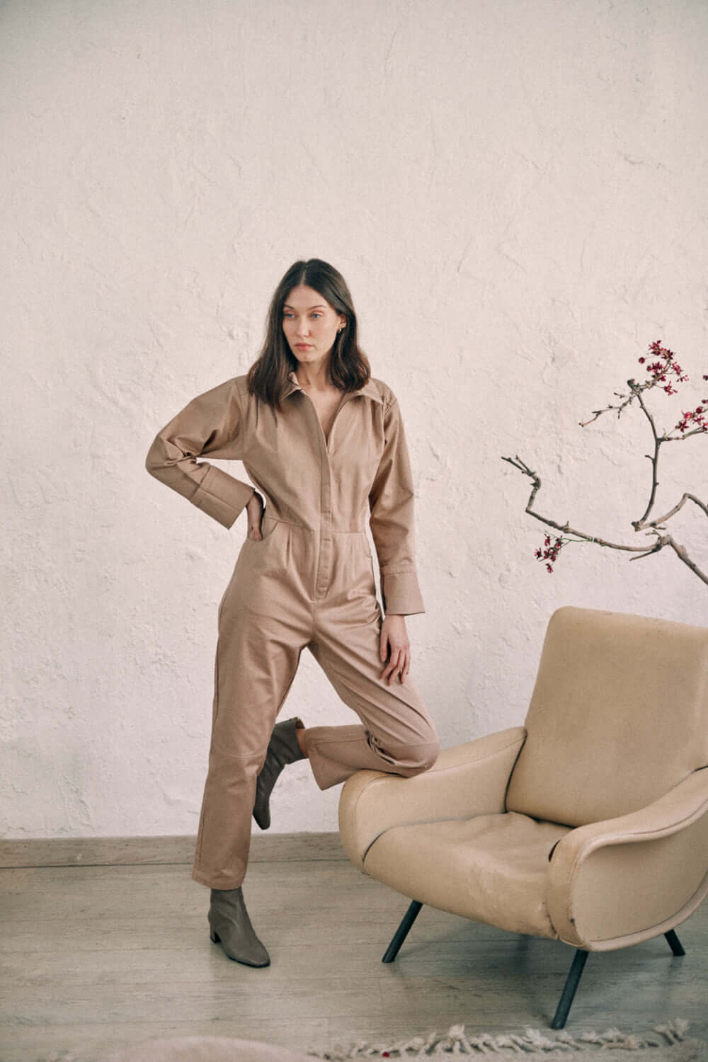 Woman wearing taupe denim boilersuit standing next to beige chair in minimalistic room.