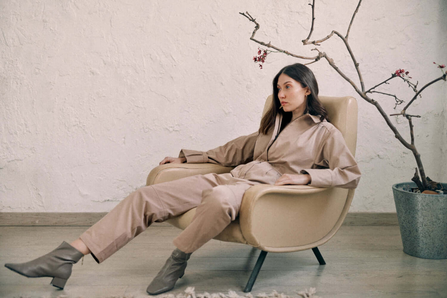 Woman in taupe denim boilersuit sitting stylishly in a chair near a minimalist decor setup.