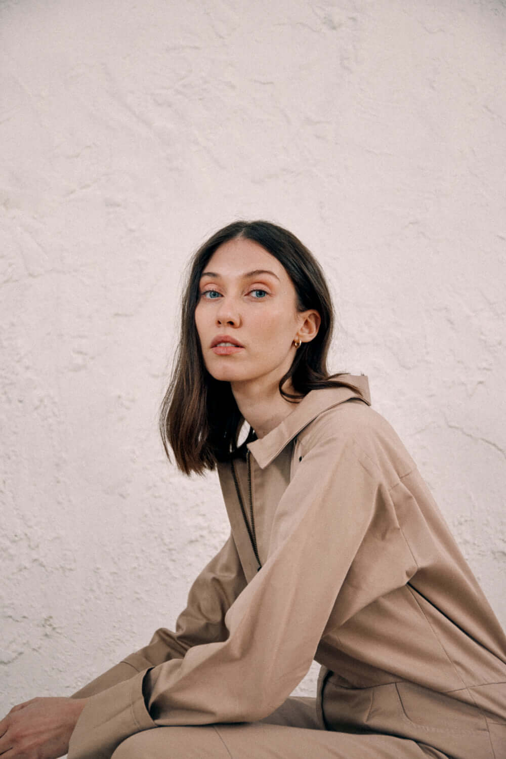 Woman wearing taupe denim boilersuit made from organic cotton, against a textured wall background.