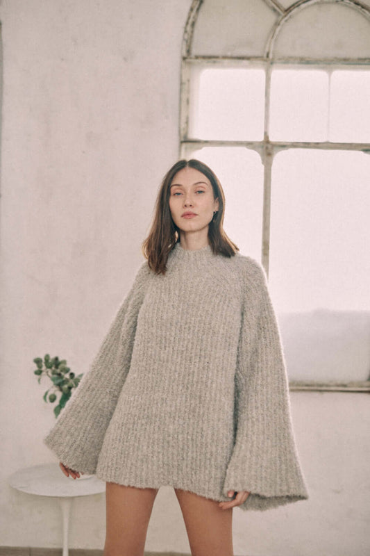 Woman wearing a grey wool tunic sweater made from 75% undyed alpaca and 25% recycled polyester, standing in a softly lit room.