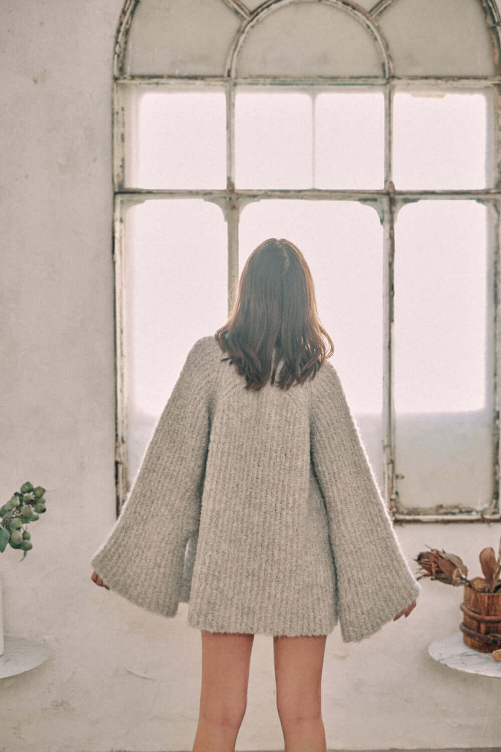 Woman wearing grey wool tunic sweater, standing by a rustic window, showcasing long sleeves and soft texture.