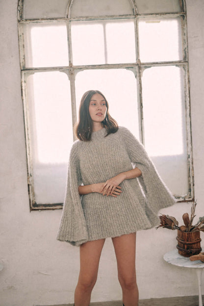 Woman wearing grey wool tunic sweater made of alpaca and recycled polyester, standing by a rustic window.